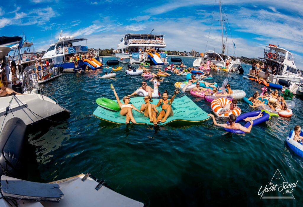 A lively scene at Yacht Social Club. Groups of people are relaxing on various colorful inflatables and boats in clear blue water. The sky is partly cloudy, and the atmosphere is cheerful and vibrant. Luxury Yacht Rentals Sydney logo features prominently in the bottom right corner.