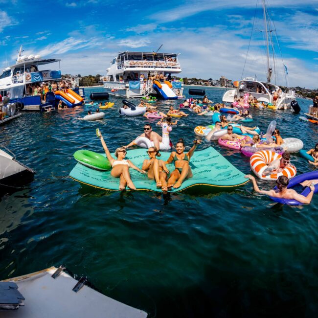 A lively scene at Yacht Social Club. Groups of people are relaxing on various colorful inflatables and boats in clear blue water. The sky is partly cloudy, and the atmosphere is cheerful and vibrant. Luxury Yacht Rentals Sydney logo features prominently in the bottom right corner.
