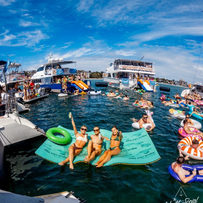 A vibrant scene of people enjoying a sunny day on the water. Various individuals are lounging on inflatable floats and boats. Three women are seated on a large floating mat, smiling, one raising a drink. Boats and other inflatables from The Yacht Social Club Event Boat Charters are visible in the background.