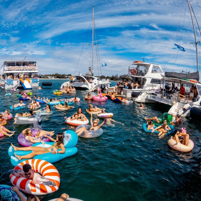 A lively marina scene with numerous people enjoying a sunny day in the water. Many individuals are on colorful inflatables of various shapes, while others are on boats around the area. A clear blue sky with scattered clouds completes the vibrant atmosphere, reminiscent of Boat Parties Sydney The Yacht Social Club.
