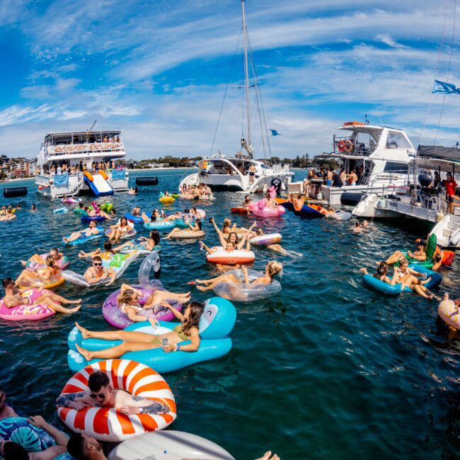 A large group of people relax on various colorful inflatable floats in the water near several docked boats and yachts on a sunny day. The sky is clear with wispy clouds, and everyone seems to be enjoying a festive atmosphere, embodying the spirit of Sydney Harbour Boat Hire The Yacht Social Club.