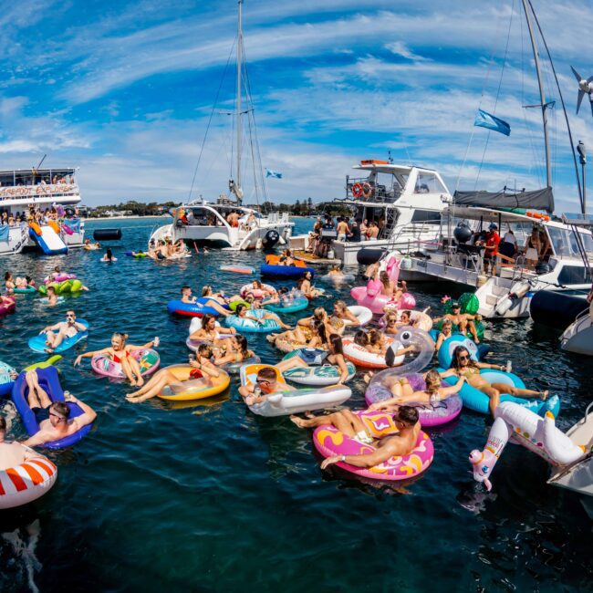 A lively scene of people enjoying a yacht party on a sunny day. Numerous individuals are floating on colorful inflatable rings and lounging in the clear blue water. Several boats and yachts from The Yacht Social Club are anchored nearby. The sky is blue with some wispy clouds, perfect for boat parties in Sydney.
