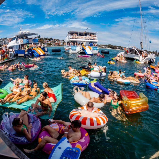 A lively scene of people enjoying a sunny day on a large body of water. Many are floating on colorful inflatable devices, including shimmery floats, pool rings, and loungers. Several boats from The Yacht Social Club are anchored nearby, with groups mingling and having fun.