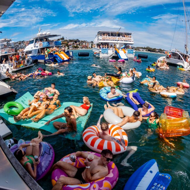 A lively scene of people enjoying a sunny day on inflatable pool floats in the water, surrounded by yachts. The Yacht Social Club hosts this vibrant gathering, with colorful floats of various shapes and sizes. Everyone appears to be relaxing, socializing, and having a good time.