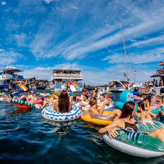 A lively gathering of people on inflatable tubes and rafts in a sunny, crowded marina. Boats and yachts from The Yacht Social Club Sydney Boat Hire form a semi-circle around the swimmers, with a clear blue sky overhead and scenic hills as a backdrop. Some people dance, enjoying the festive atmosphere.