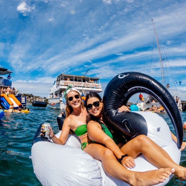 Two women are enjoying a sunny day on an inflatable swan float in an ocean area, smiling and wearing sunglasses. The backdrop of boats and a clear blue sky with scattered clouds adds to the lively, festive atmosphere, reminiscent of a Boat Party hosted by The Yacht Social Club.