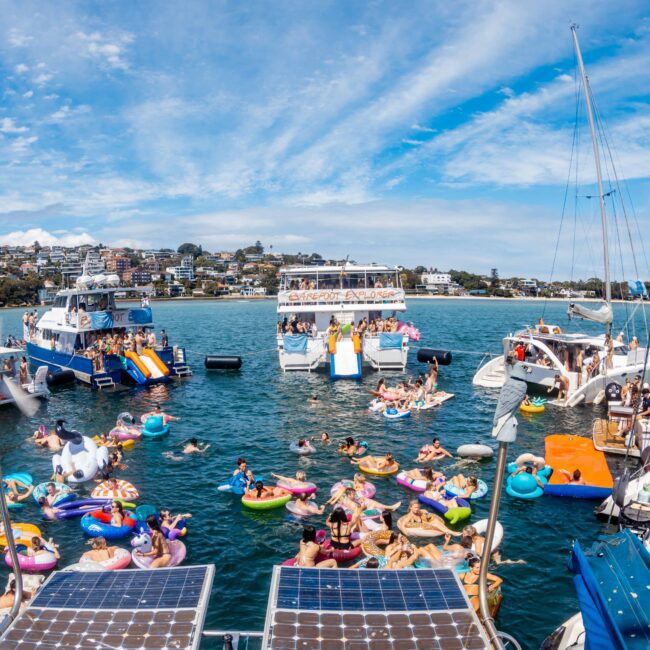 A lively scene on a sunny day at a bay with multiple anchored boats from The Yacht Social Club Sydney Boat Hire. People are enjoying themselves on floaties and swimming in the water. Surrounding hills are dotted with houses, creating a festive atmosphere with clear skies and calm blue waters.