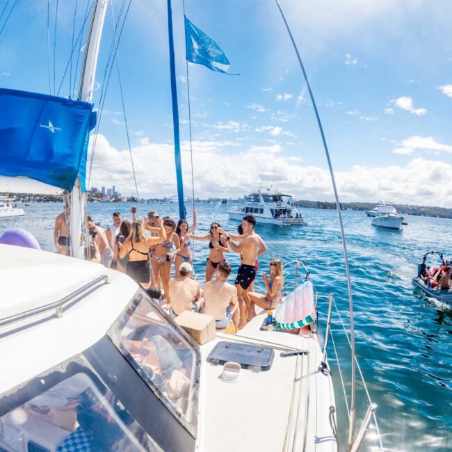 A group of people in swimwear gather on the deck of a sailboat, enjoying a sunny day on the water in Sydney Harbour. Other boats are visible in the background, along with a clear blue sky and some fluffy clouds. A logo at the bottom right reads "The Yacht Social Club Sydney Boat Hire.