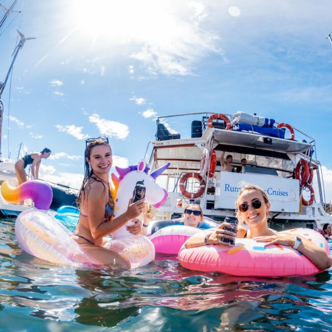 People are enjoying a sunny day on the water, floating on colorful inflatable rings near a docked boat. They are smiling, holding drinks, and having fun under a bright, clear sky. The boat in the background has a visible name along with the logo "Boat Parties Sydney The Yacht Social Club.