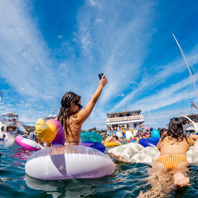 A woman raises her phone while floating on an inflatable toy in a body of water, surrounded by others on various floaties. Nearby boats and yachts from Sydney Harbour Boat Hire The Yacht Social Club are docked, and the sky is bright and blue with scattered clouds.