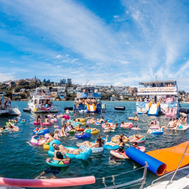 A lively scene from The Yacht Social Club Event Boat Charters is depicted, with numerous people floating on colorful inflatable rings between several yachts. The sky is clear with scattered clouds, and a waterslide is visible on one of the boats. The backdrop includes a hilly, wooded shoreline.