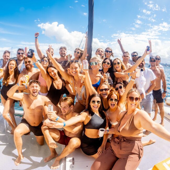 A large group of people enjoying a sunny day on a boat. They are smiling, cheering, and posing for the camera, dressed in swimwear and casual summer outfits. The bright blue sky and water create a vibrant, festive atmosphere at The Yacht Social Club Sydney Boat Hire.