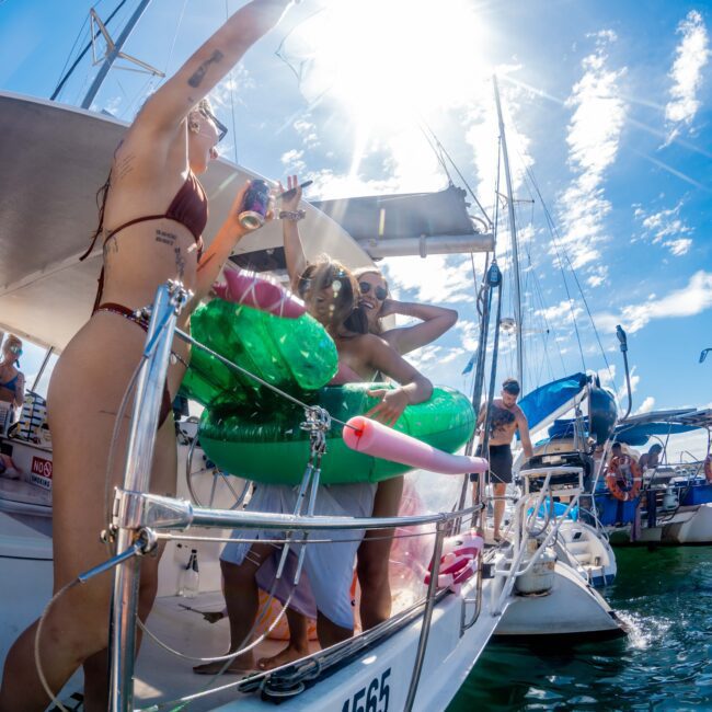 A lively group enjoys a sunny day on a boat, with several people in swimsuits dancing and holding drinks. One person sprays a mist of water or champagne into the air. Inflatable toys and pool floats add to the festive atmosphere, embodying the spirit of Boat Parties Sydney The Yacht Social Club.