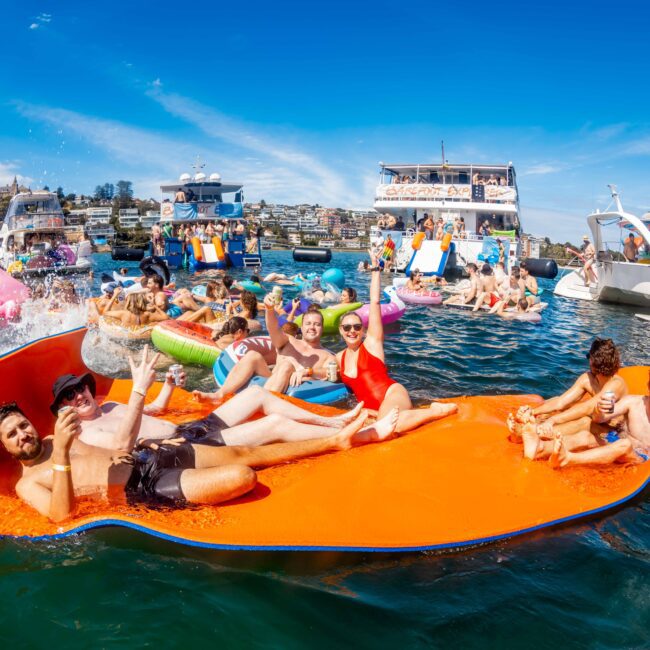 A group of people enjoying a sunny day on the water, lounging on an orange inflatable mat, and socializing on various boats and floating toys around them. The background features clear blue skies and more watercraft. Logo text reads "The Yacht Social Club: Sydney Harbour Boat Hire.