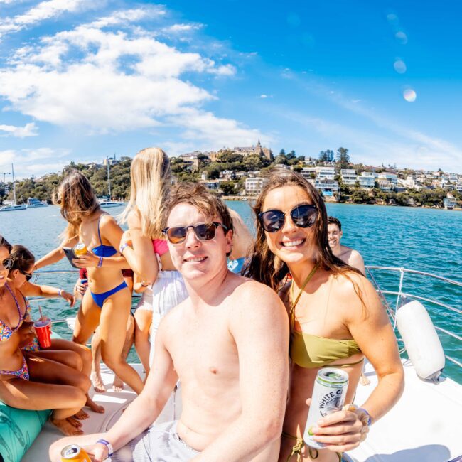 A group of people are enjoying a sunny day on a boat from The Yacht Social Club Sydney Boat Hire. Some are seated while others are standing, all wearing swimwear and holding drinks. The background features a clear sky, calm waters, and a seaside town with houses on a hill.