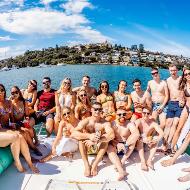 A large group of people in swimsuits and summer attire are smiling and posing for a photo on the deck of a boat. The Yacht Social Club Sydney Boat Hire is on a large body of water, with a coastline and houses visible in the background under a bright blue sky.