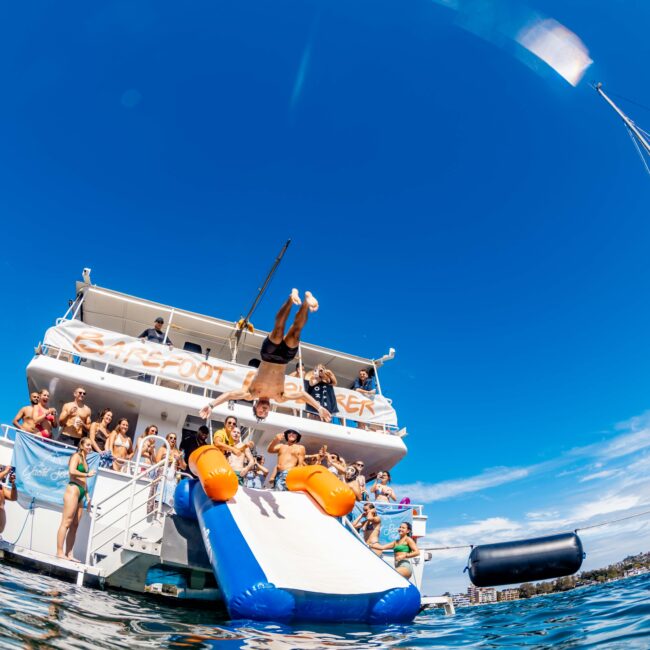 A man flips off an inflatable slide on a boat filled with people, while others watch from the deck. The boat is on a calm body of water under a clear blue sky. A sailboat mast is visible to the right, and several people are enjoying a sunny day at The Yacht Social Club Sydney Boat Hire.