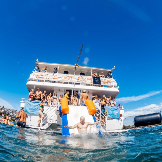 A crowded party boat named "Cabana Boat Explorer" floats on the water. People are enjoying themselves, with a man sliding down a water slide into the sea. The sky is clear and blue, adding to the festive atmosphere. Known for its lively and joyful scenes, it's perfect for boat parties with Sydney Harbour Boat Hire The Yacht Social Club.