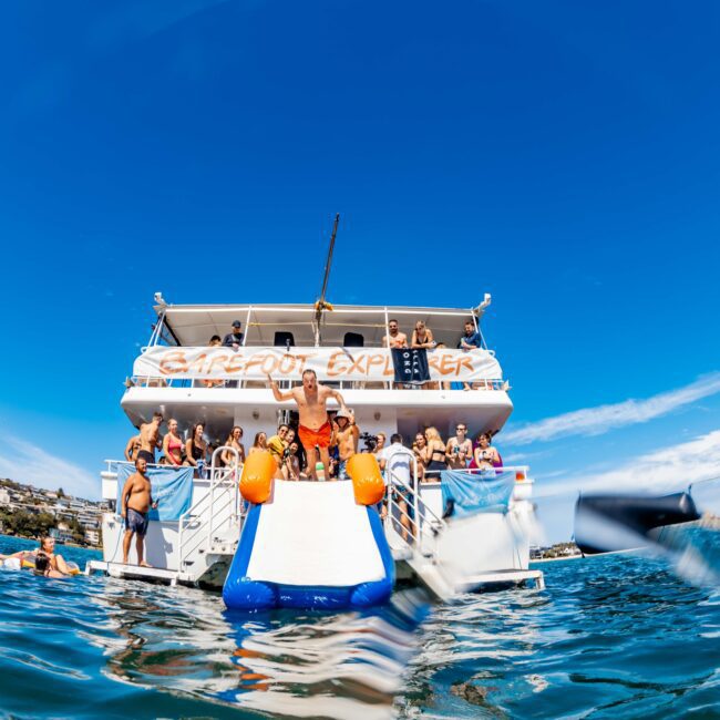 A lively group of people are aboard a large boat named "Barefoot Explorer," docked on clear blue waters. Passengers are either on the deck or enjoying a water slide splashing into the sea. The sky is bright and clear, indicating a sunny day—perfect for Luxury Yacht Rentals Sydney.