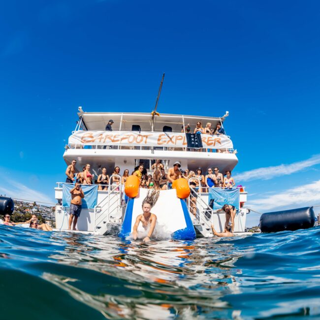 A vibrant scene of a boat party on the ocean, featuring passengers sliding into the water from two slides at the back. The boat, chartered by The Yacht Social Club Sydney Boat Hire, is adorned with a "CareOut Experience" banner. The weather is sunny with a clear blue sky and calm waters.