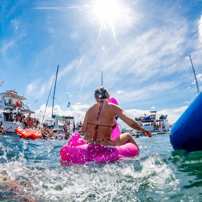 People enjoying a sunny day at a yacht event in Sydney. The image shows boats anchored close together with partygoers swimming and relaxing on colorful inflatables. Boats and inflatable toys are prominent at The Yacht Social Club Event Boat Charters, with a clear blue sky and the sun shining brightly.