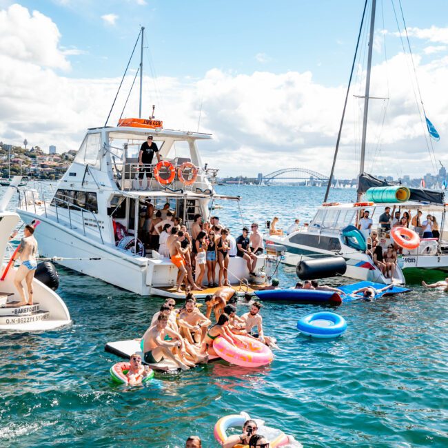A lively scene of people enjoying a sunny day on the water. Several are on a boat, others float on colorful inflatable rings and toys in the water. A bridge is visible in the background. The sky is blue with scattered clouds. The atmosphere at The Yacht Social Club Event Boat Charters appears joyous and festive.