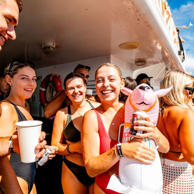 A group of people in swimsuits enjoy a sunny day on a boat. A smiling woman holds a pink inflatable kangaroo while others around her hold drinks and chat. The Yacht Social Club offers perfect boat parties in Sydney on the water, with clear blue skies in the background.