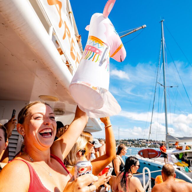 A person in a red swimsuit and wristband smiles while holding up a pool float shaped like a unicorn. They are standing on a boat filled with other people under a sunny blue sky. Another boat and a dock with more people are visible in the background, embodying the spirit of Boat Parties Sydney The Yacht Social Club.