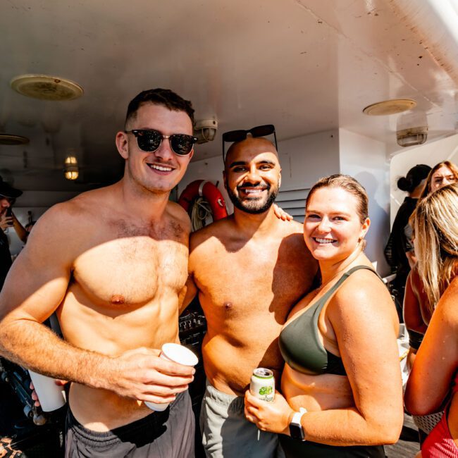 A group of people are enjoying a sunny day on a boat from The Yacht Social Club Sydney Boat Hire. Three individuals in the forefront are smiling at the camera, two men without shirts, and a woman in a swimsuit. Other passengers and the boat's structure are visible in the background.