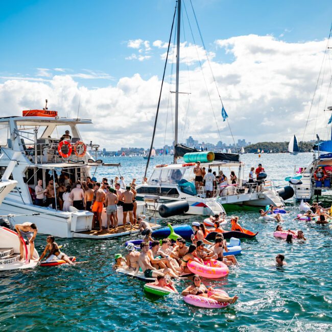 A lively scene at a boat party on a sunny day. Numerous people are in the water on colorful inflatable floats, socializing and enjoying the weather. Several boats are anchored nearby, filled with more partygoers from The Yacht Social Club Sydney Boat Hire. The sky is partly cloudy with the city skyline in the distance.