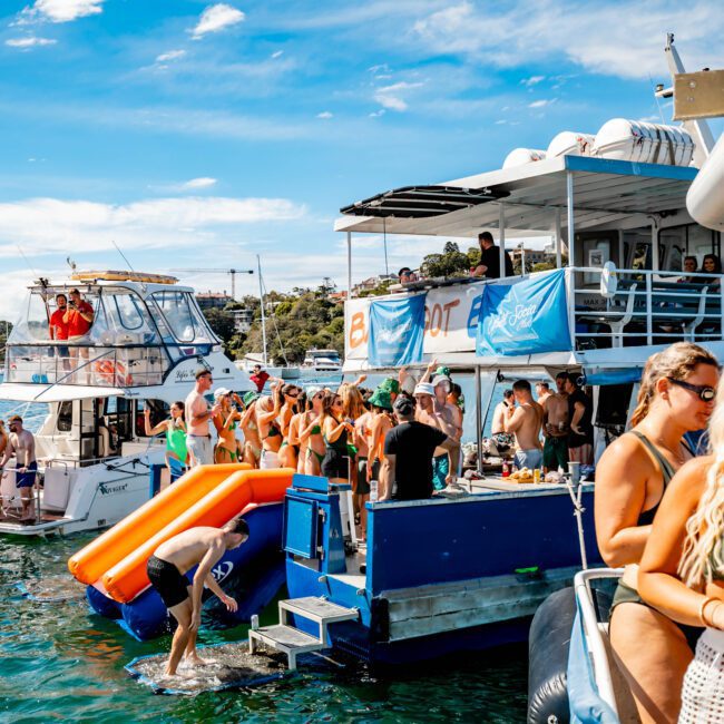 A lively Boat Parties Sydney The Yacht Social Club event with people swimming, relaxing, and socializing. The scene features two boats anchored close to each other with an inflatable slide connecting them. Attendees are enjoying the sunny weather, with blue skies and greenery in the background.