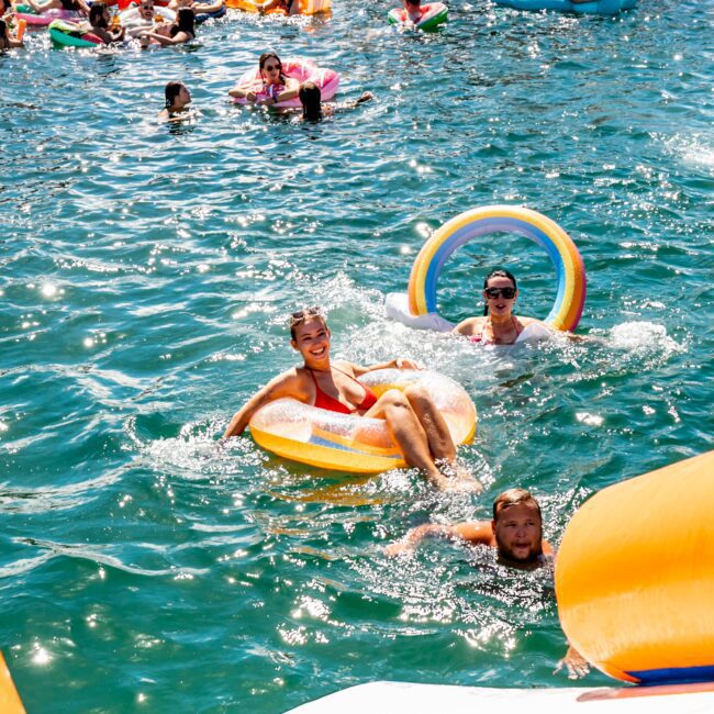 A large group of people enjoy a sunny day at the beach, floating in the water on colorful inflatable rings. Several individuals are near a docked boat labeled "Rum Runner Cruises." The scene is lively with swimmers, and the water is clear and inviting. Consider Luxury Yacht Rentals Sydney for an elevated experience.