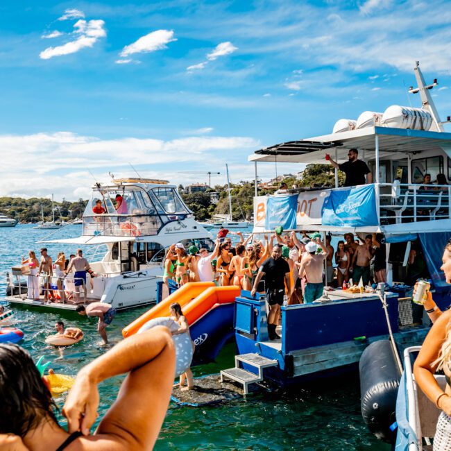 A lively boat party on a sunny day features several boats anchored together with numerous people on board and in the water. Attendees of The Yacht Social Club enjoy music, swimming, and socializing. The blue skies and calm water create a festive and vibrant atmosphere perfect for such events.