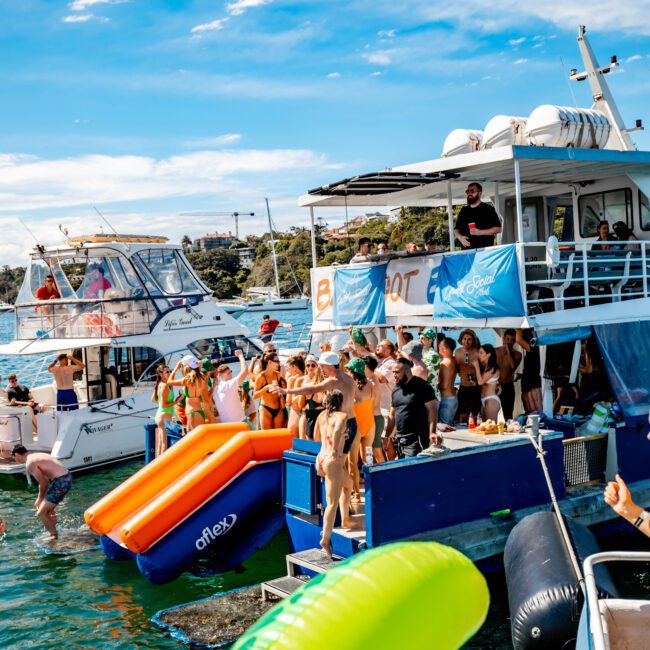 A vibrant scene of people enjoying a sunny day on boats. One boat is full of people in swimwear, some wearing hats and sunglasses. Inflatable items like a green floaty and an orange slide are visible. The blue sky and water create a cheerful atmosphere, perfect for Boat Parties Sydney The Yacht Social Club.