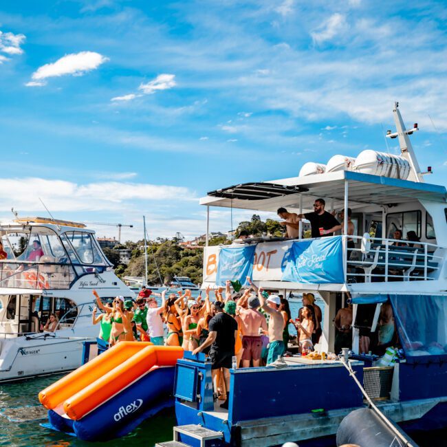 A group of people enjoying a party on a yacht in sunny weather. The Yacht Social Club Event Boat Charters decorated the yacht with blue banners and attached an orange slide leading into the water. Several individuals are dancing, socializing, and taking in the scenic views of the surrounding water and shoreline.