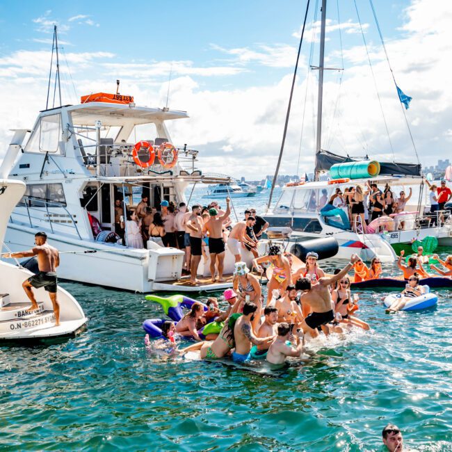 A lively scene of people enjoying a boat party hosted by The Yacht Social Club Sydney Boat Hire. Several individuals are on the boat, while others float on inflatables under a clear sky. Nearby, anchored boats add to the festive atmosphere as everyone delights in the sun and water activities.