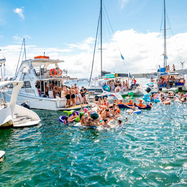 A lively scene of people enjoying a sunny day on the water. Several boats and yachts from The Yacht Social Club Sydney Boat Hire are anchored close together as people swim and float on colorful inflatables in the turquoise water. The sky is clear with a few clouds, and the atmosphere is festive.