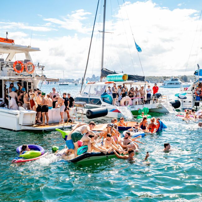 A lively boat party is in full swing with many people enjoying themselves on and around several yachts. Some are in the water on inflatables, while others dance and socialize on the boats. The Yacht Social Club Event Boat Charters adds a touch of elegance to the festive atmosphere under a bright sky with scattered clouds.