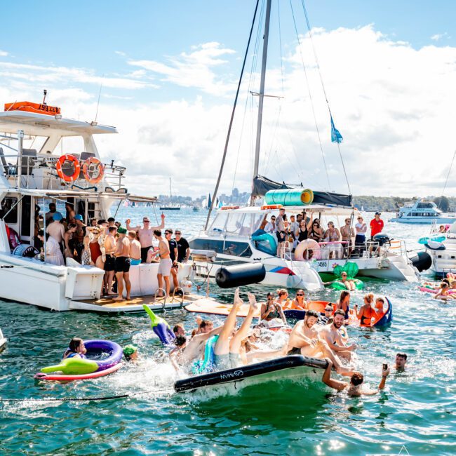 A lively scene of people enjoying a sunny day on the water, with multiple boats docked close together. Groups are swimming, relaxing on floats, and socializing on the boats, creating a festive atmosphere. The Yacht Social Club Sydney Boat Hire ensures the sky is clear and the water sparkling for all.