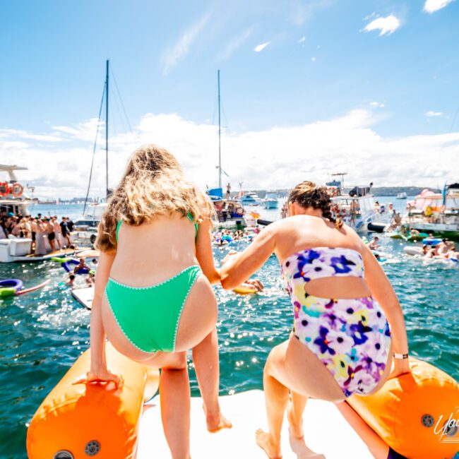 Two girls in swimsuits are seen from behind as they prepare to jump into a crowded lake from an inflatable float. Both are holding hands while many others are swimming, floating, or on boats nearby on a sunny day, perhaps dreaming of Luxury Yacht Rentals Sydney.