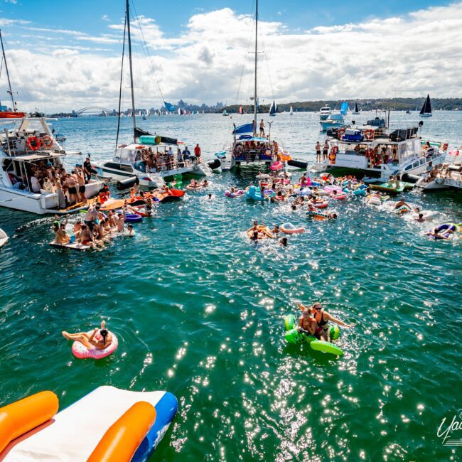 A lively scene of people enjoying a party on a sunny day in a bay. Several boats from The Yacht Social Club are anchored close together with people socializing on board and in the water, floating on inflatables. The background includes a clear blue sky with some clouds and a cityscape.