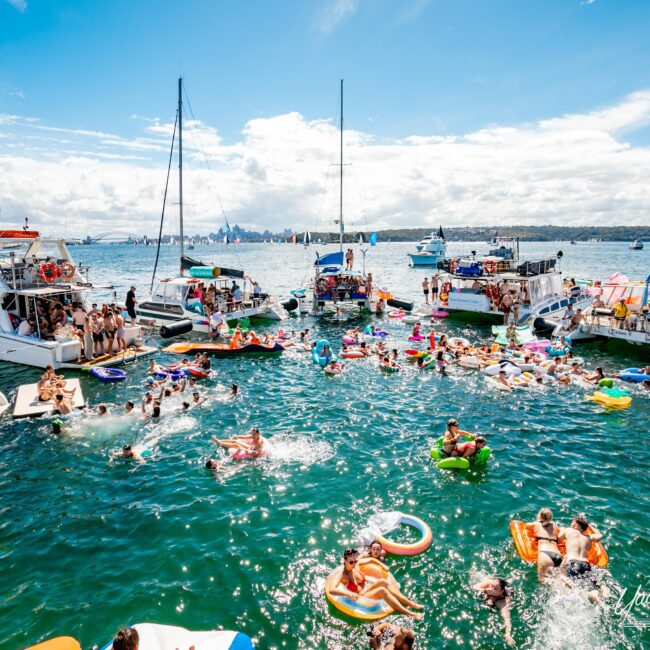 A lively scene of people swimming and lounging on colorful inflatables in a sunlit body of water surrounded by numerous anchored boats. The sky is clear with a few clouds, and the distant shoreline is visible. The image exudes a festive, vibrant atmosphere typical of Sydney Harbour Boat Hire The Yacht Social Club events.