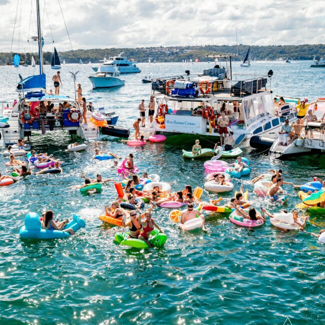 A vibrant scene of a yacht party by The Yacht Social Club where people are socializing and enjoying the water. Many are floating on colorful inflatable rafts in a busy harbor filled with boats. The atmosphere is festive with blue skies and calm waters, showcasing a lively gathering perfect for boat parties in Sydney.