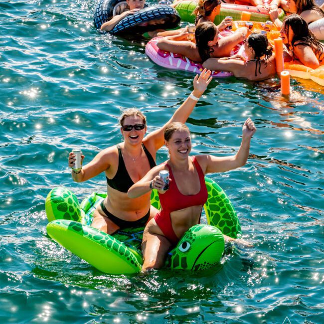Two women in swimsuits joyfully float on a green inflatable turtle in a body of water. They are surrounded by other people on various inflatables, some with drinks in hand. The scene is festive, depicting a lively social gathering akin to The Yacht Social Club Sydney Boat Hire events.