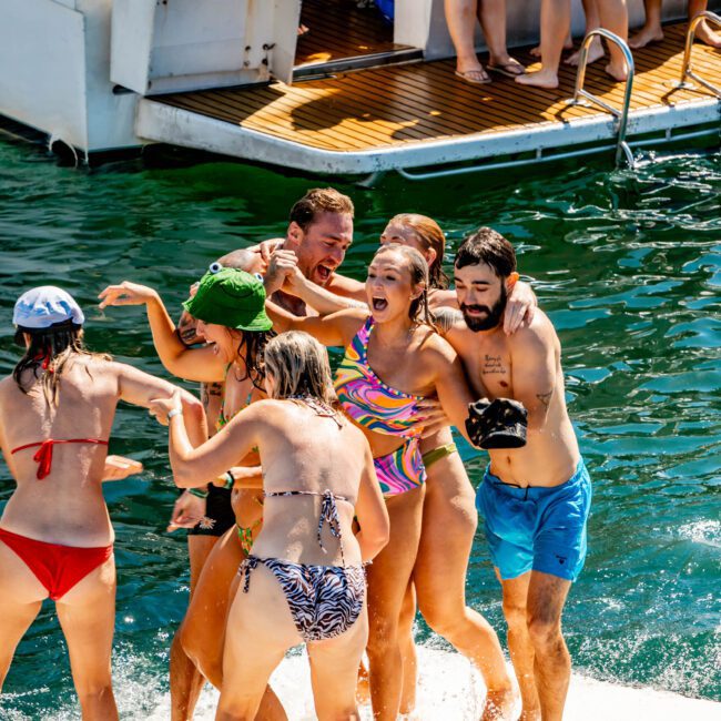 A group of people in swimwear enjoy a fun, lively moment on a boat's deck and in the water. Some are embracing and laughing, while others stand nearby. The greenish-blue water adds to the charm as more people are seen in the background, experiencing Boat Parties Sydney with The Yacht Social Club.