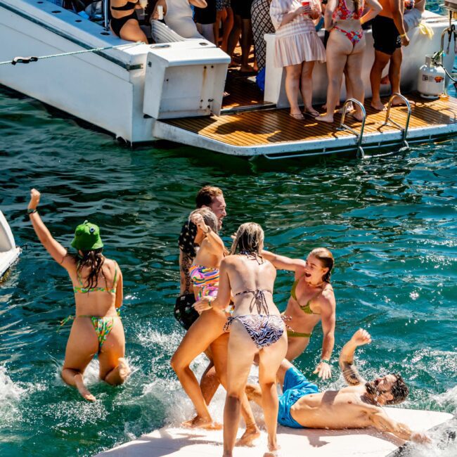 A lively group of people are enjoying a sunny day with The Yacht Social Club Event Boat Charters. Some are dancing and socializing on the boat's deck, while others are playing and splashing in the water near a floating platform. Everyone appears to be in swimsuits, having fun.