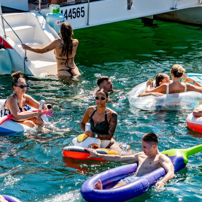 A group of people enjoying a sunny day in the water near a boat. Some are floating on colorful inflatables, while others are swimming or sitting on the edge of the boat. The lively scene, hosted by The Yacht Social Club Sydney Boat Hire, features clear blue-green water and bright sunlight.