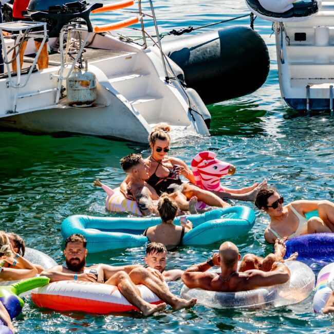 A group of people enjoy floating on inflatable rafts in the calm, inviting water near docked boats. Some rafts are plain, while others are shaped like animals. A person on a boat grills food in the background, part of The Yacht Social Club Event Boat Charters’ vibrant atmosphere.