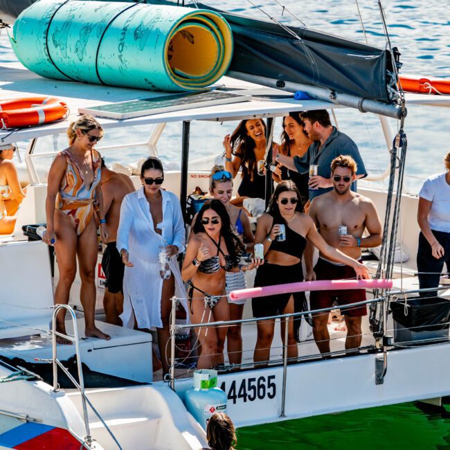 A group of people enjoy a sunny day on a sailboat in Sydney Harbour, holding drinks and smiling. One person is in the water by the boat's ladder. Pool floats are attached to the sailboat's roof. The boat is on a calm body of water, creating a fun and relaxed atmosphere, courtesy of The Yacht Social Club.