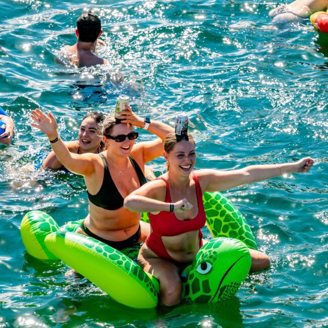 Two women in swimsuits laugh and pose on an inflatable green turtle in a sunlit body of water. One woman balances a drink can on her head while the other flexes her arm. Other swimmers and inflatable toys are visible in the background, giving it a relaxed vibe reminiscent of The Yacht Social Club Event Boat Charters.
